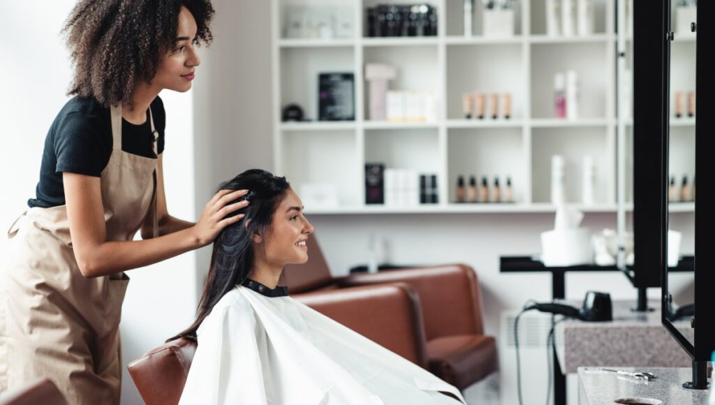 A hairdresser and her client looking at a hair salon mirror. Behind them is a shelf full of organized beauty products.