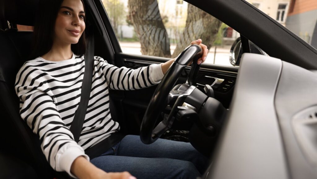 A brunette woman in a black and white striped shirt using one hand to shift gears while driving a car.