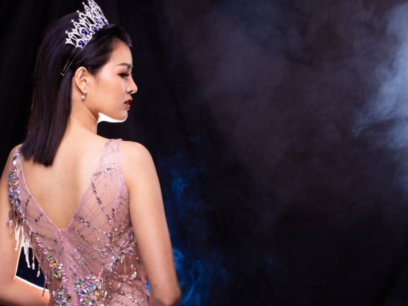 A woman in a pink pageant dress and tall crown with blue gems looks over her shoulder against a black, smoky background.