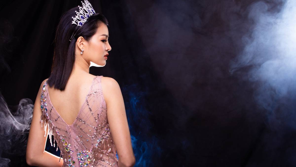 A woman in a pink pageant dress and tall crown with blue gems looks over her shoulder against a black, smoky background.