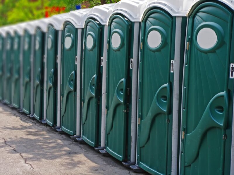 A long row of forest green portable restroom units on black pavement outside on a sunny day.