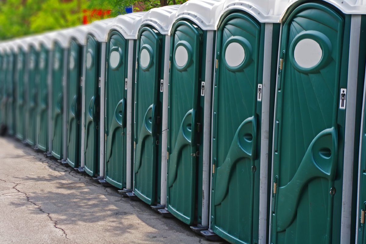 A long row of forest green portable restroom units on black pavement outside on a sunny day.