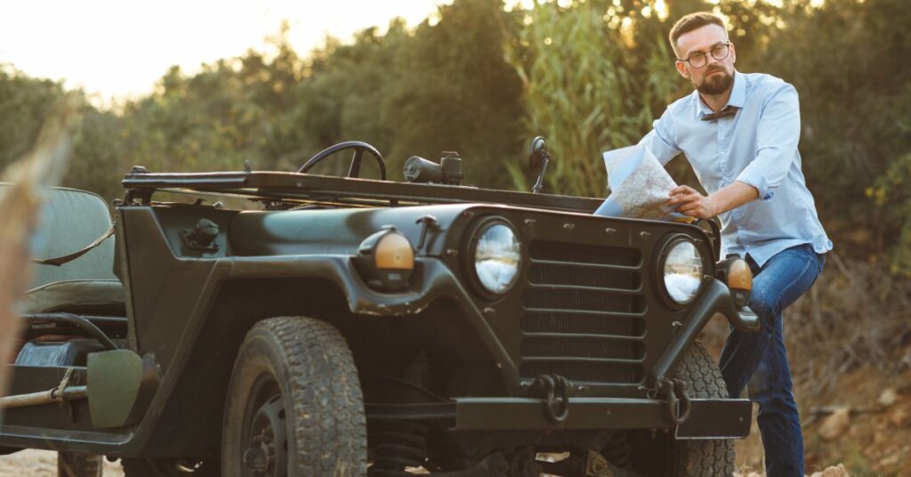 A young man wearing a blue shirt and jeans reading a map while leaning against a dark green vintage jeep.