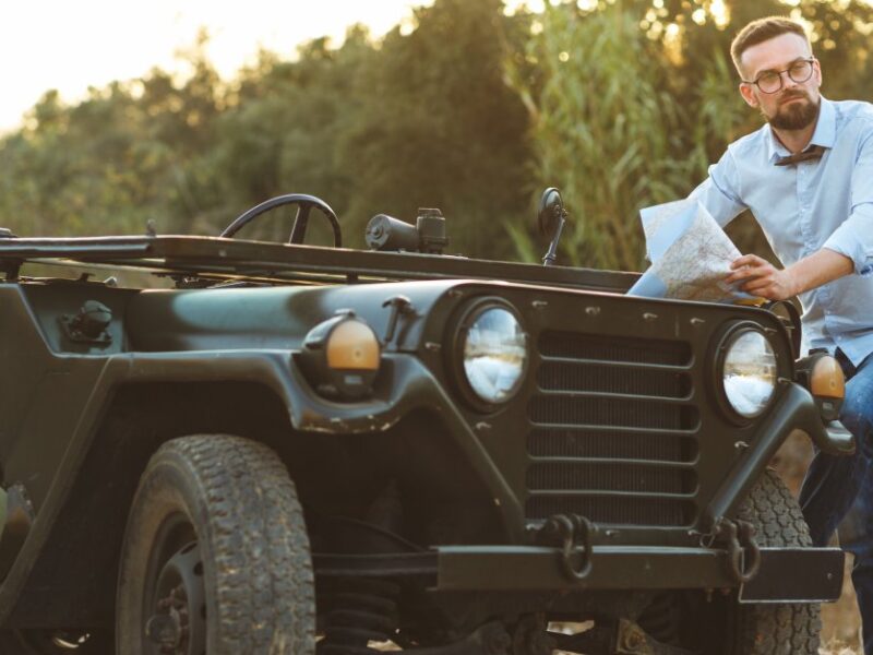 A young man wearing a blue shirt and jeans reading a map while leaning against a dark green vintage jeep.