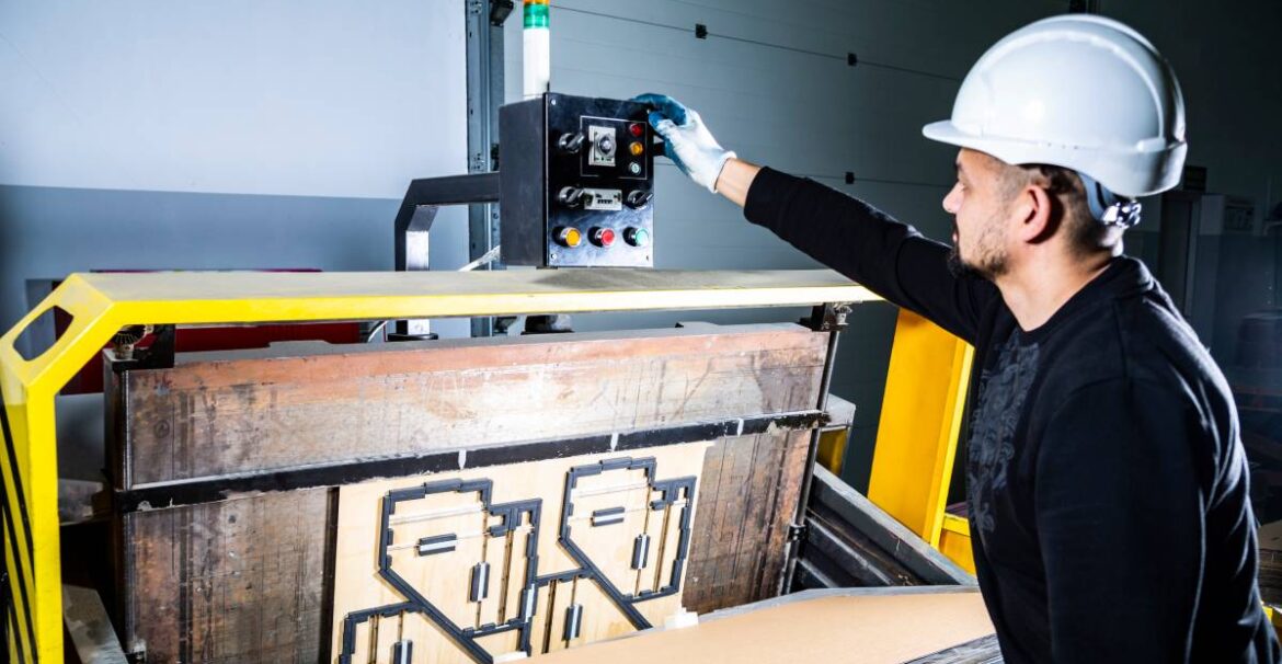 A man working as a packaging machine operator presses a button on an industrial die cutting machine for cardboard.