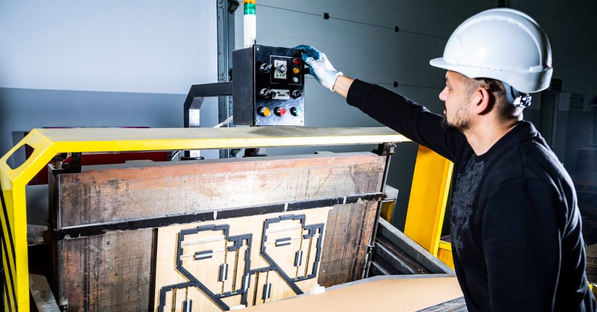 A man working as a packaging machine operator presses a button on an industrial die cutting machine for cardboard.