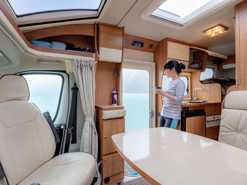 The interior of a clean and organized RV. A young woman is holding a mug and standing by the open door.