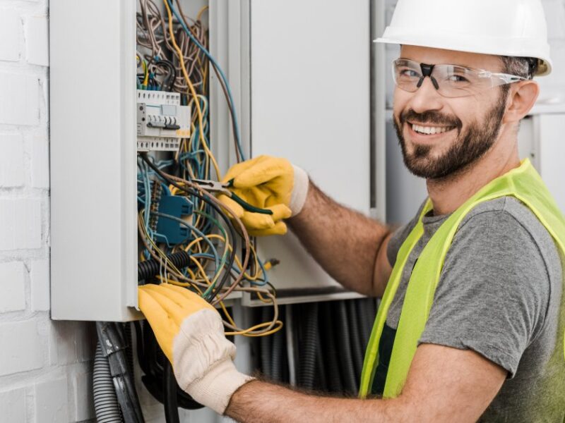 An electrician wearing a white hard hat and a yellow safety vest smiles as he works on an electrical box.