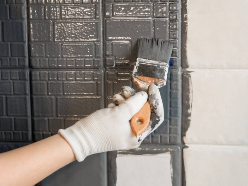 A person's hand wearing a white glove and holding a paintbrush as they apply gray paint to small white wall tiles.