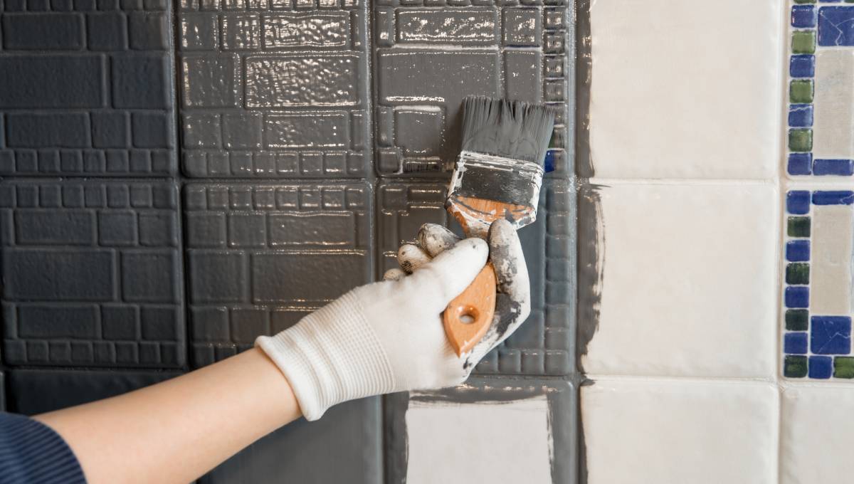 A person's hand wearing a white glove and holding a paintbrush as they apply gray paint to small white wall tiles.