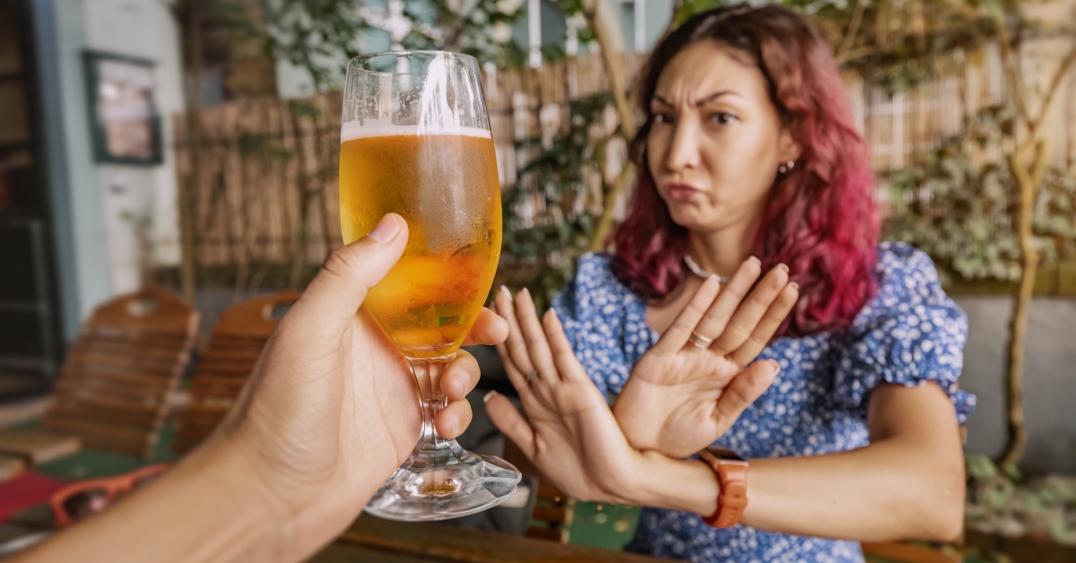 A hand holds a glass of beer out to a woman who has her arms crossed in an X infront of her.