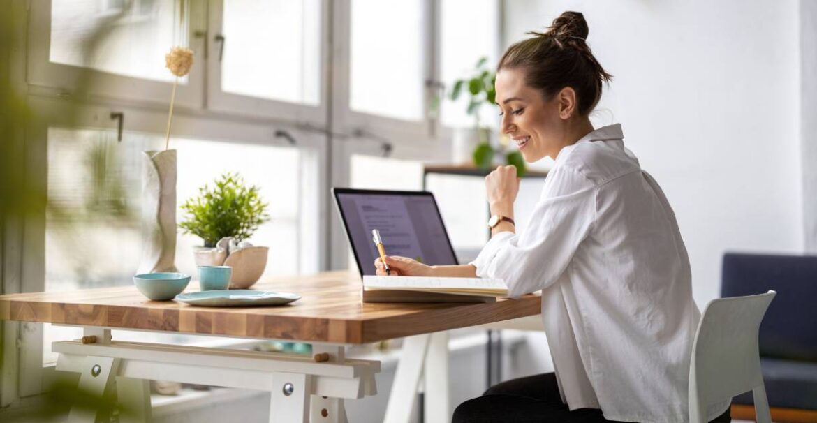 A woman works in her home office. She has a notebook and laptop open on her desk, surrounded by decorations.