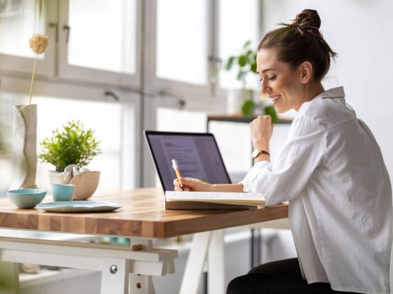 A woman works in her home office. She has a notebook and laptop open on her desk, surrounded by decorations.