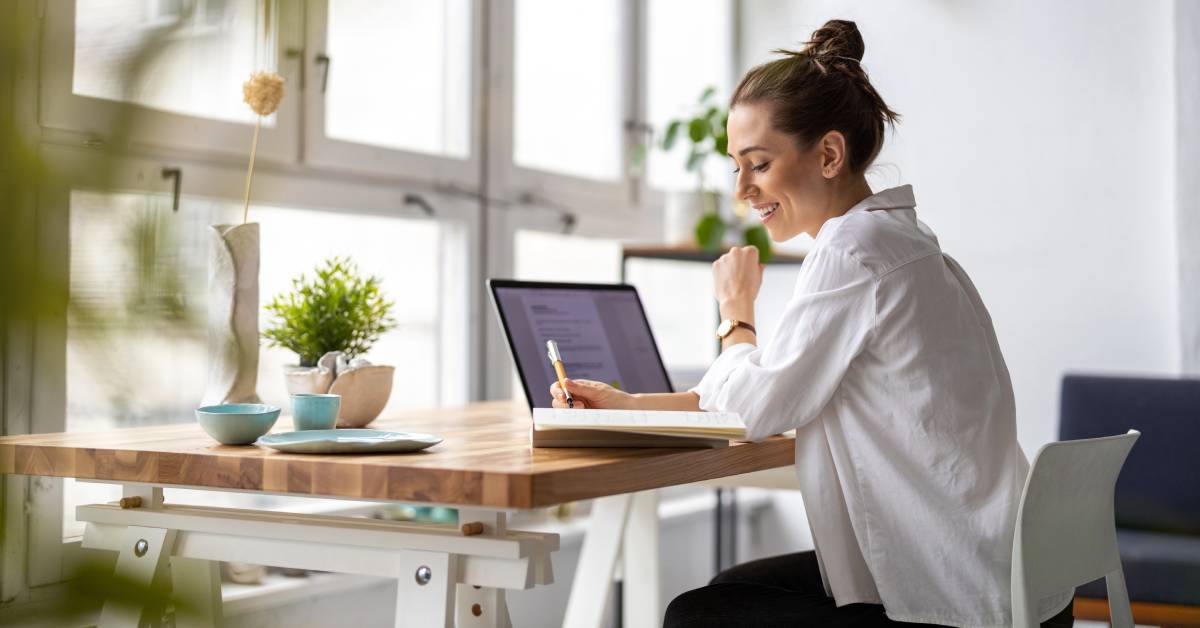 A woman works in her home office. She has a notebook and laptop open on her desk, surrounded by decorations.