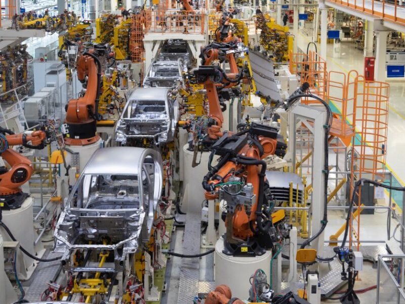 Partially-built silver cars on a conveyer belt with orange machines adding to them in a factory.