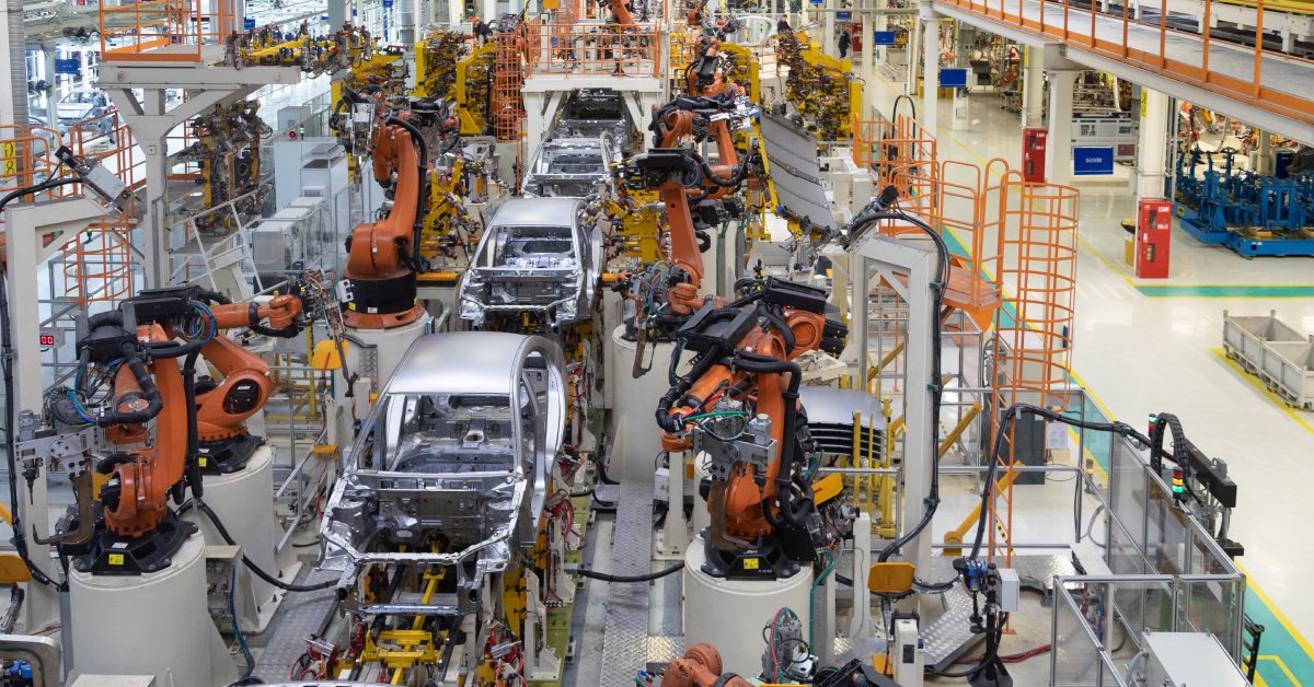 Partially-built silver cars on a conveyer belt with orange machines adding to them in a factory.