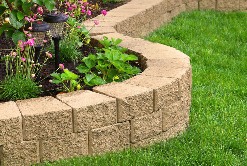 A low wall made of bricks that encircles a garden with lanterns and flowers next to a lawn with perfectly manicured grass.