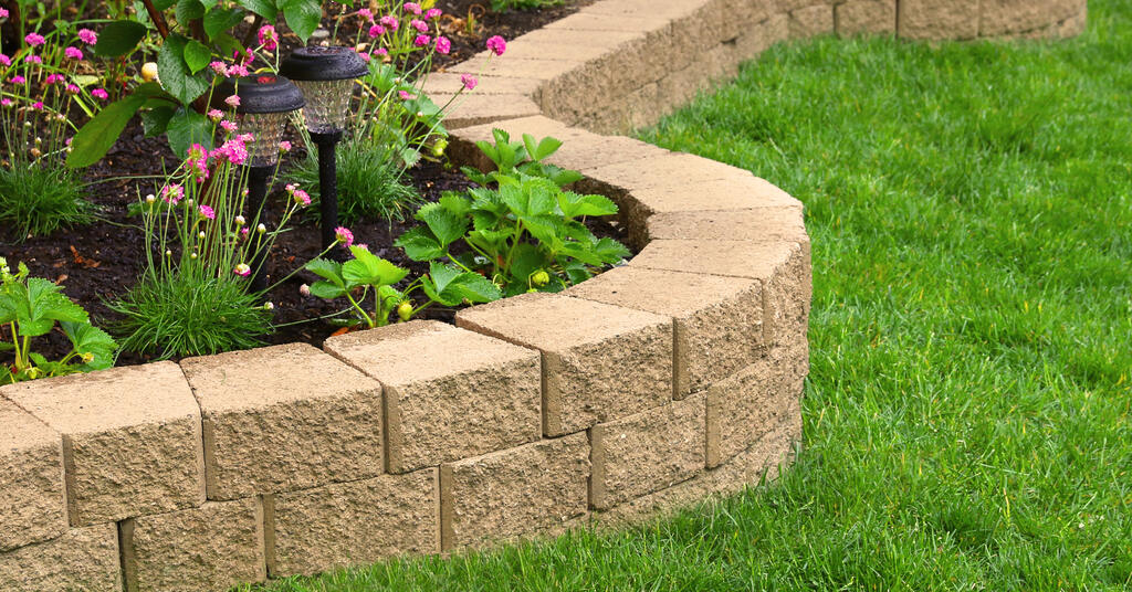 A low wall made of bricks that encircles a garden with lanterns and flowers next to a lawn with perfectly manicured grass.