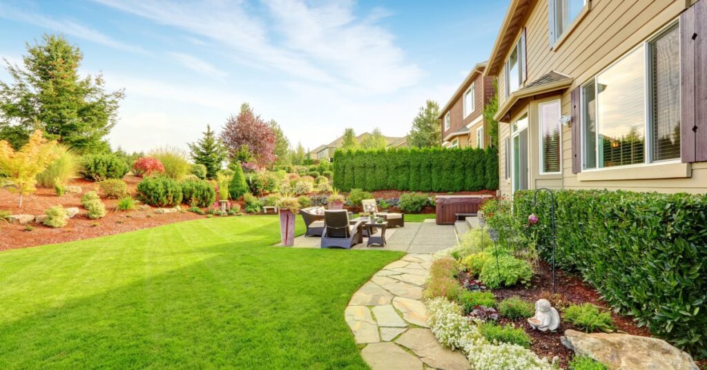 A backyard next to a house with beige siding. The yard has a patio with chairs on it and a garden.