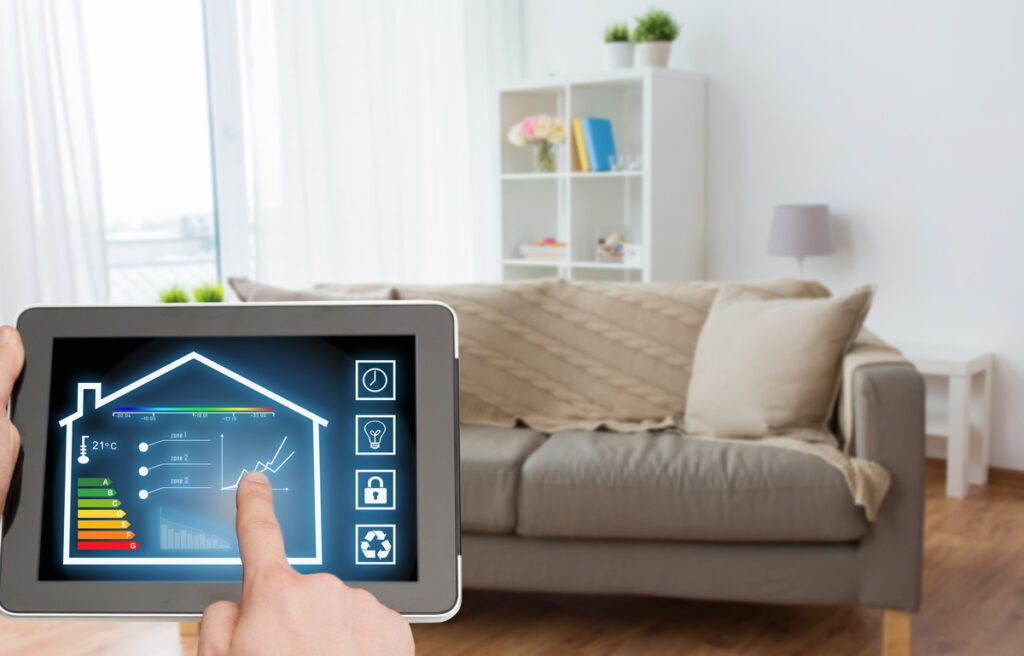 A person holding a tablet operating a smart home app while standing in their living room in front of a couch.