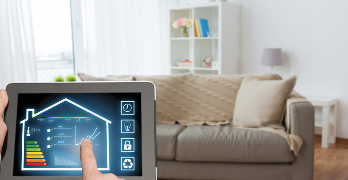 A person holding a tablet operating a smart home app while standing in their living room in front of a couch.