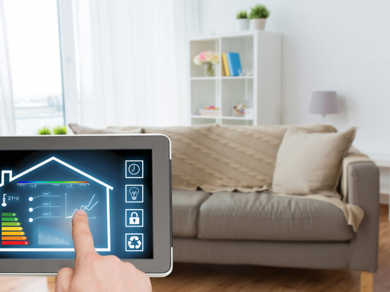 A person holding a tablet operating a smart home app while standing in their living room in front of a couch.