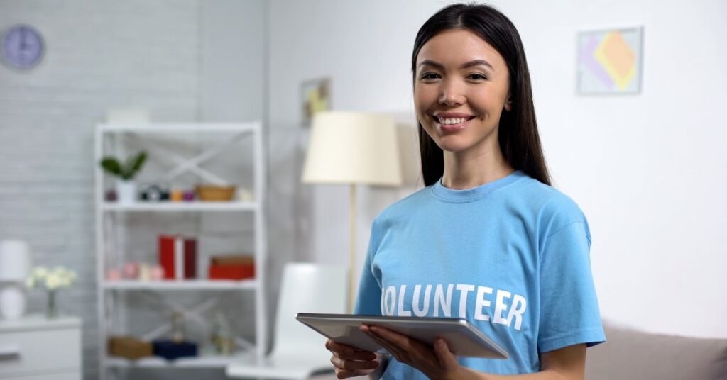 A woman is wearing a blue t-shirt that says "Volunteer" in white letters. She is smiling and holding a tablet.