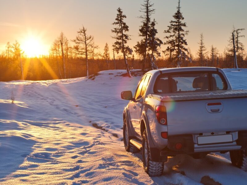 A gray pickup truck is parked on a snowy offroad trail. In the distance are evergreens and a rising sun.