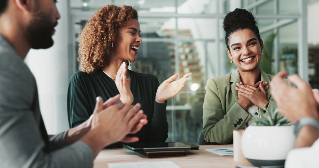 In a business meeting, employees around a table clap for a woman smiling and holding her hands to her chest.