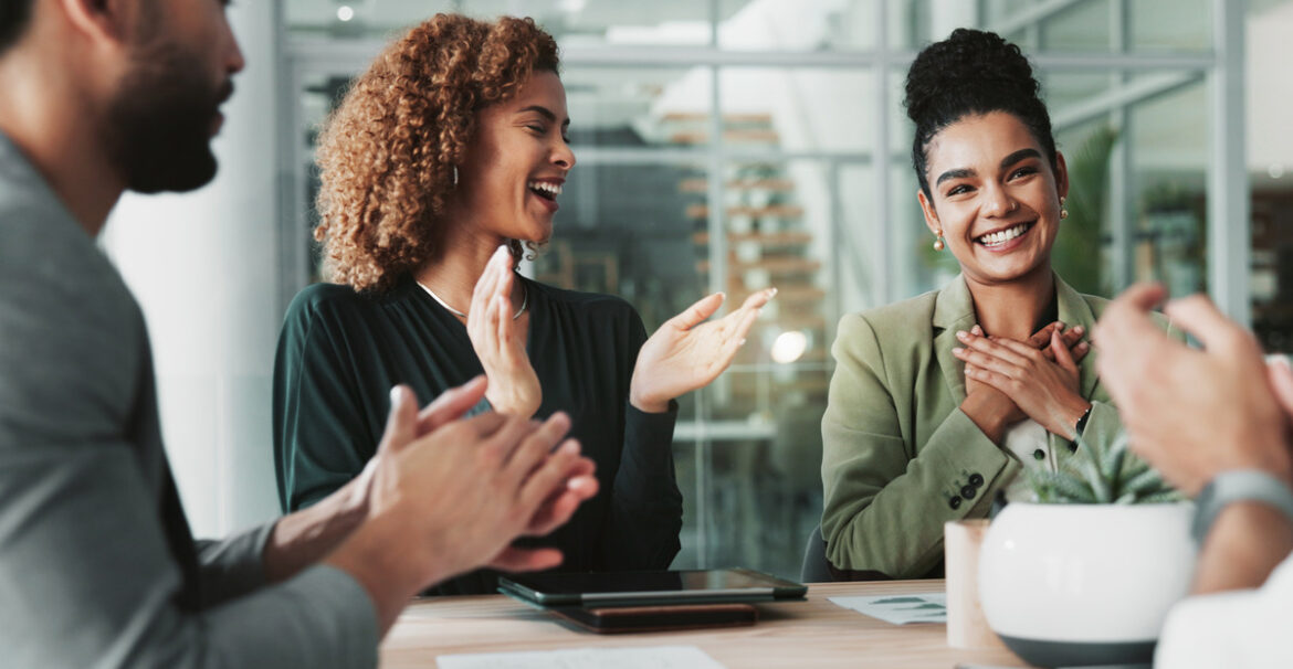 In a business meeting, employees around a table clap for a woman smiling and holding her hands to her chest.