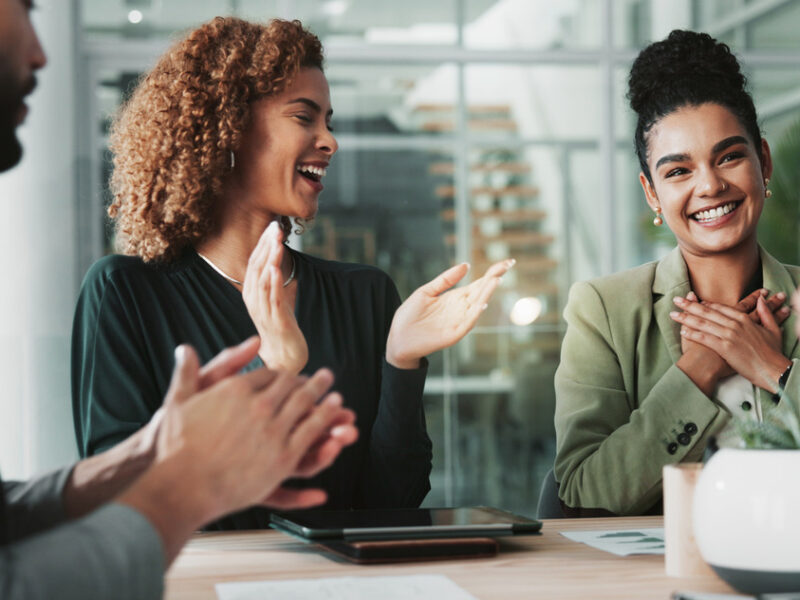 In a business meeting, employees around a table clap for a woman smiling and holding her hands to her chest.