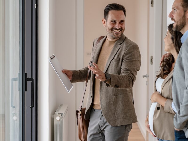 A pregnant woman and man standing beside her walk through a building guided by a real estate agent holding a clipboard.