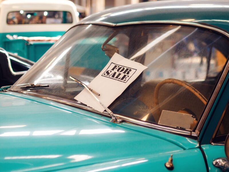 A teal vintage car with a "For Sale" sign under the windshield wiper. It is displayed indoors with other vehicles.