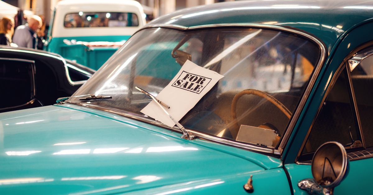 A teal vintage car with a "For Sale" sign under the windshield wiper. It is displayed indoors with other vehicles.