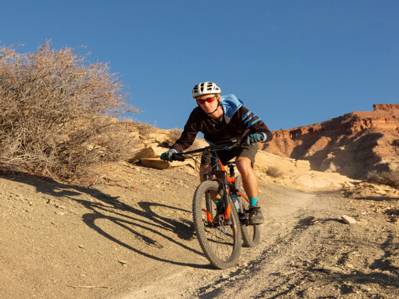 A person riding a bike through a desert landscape. He is wearing a white helmet, black gloves, and orange sunglasses.