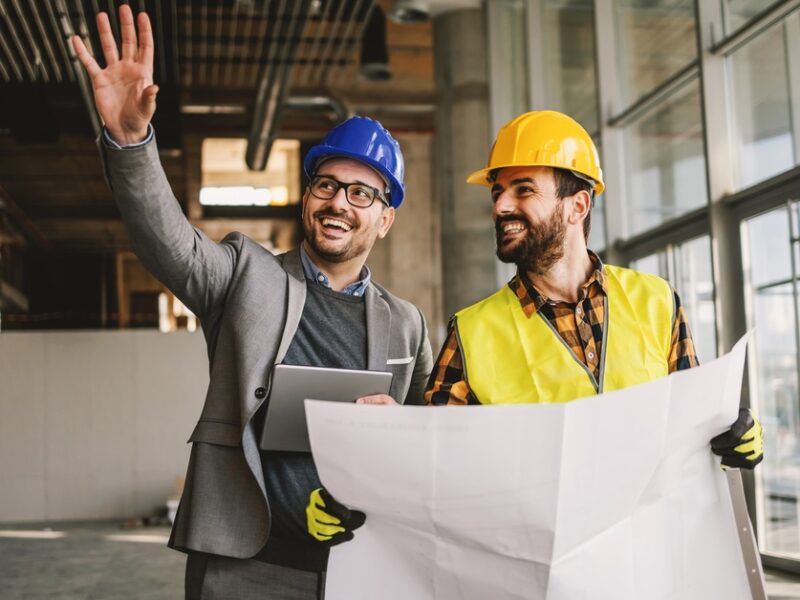 A man in a suit and hard hat gestures while standing beside a worker in a safety vest holding large architectural plans.