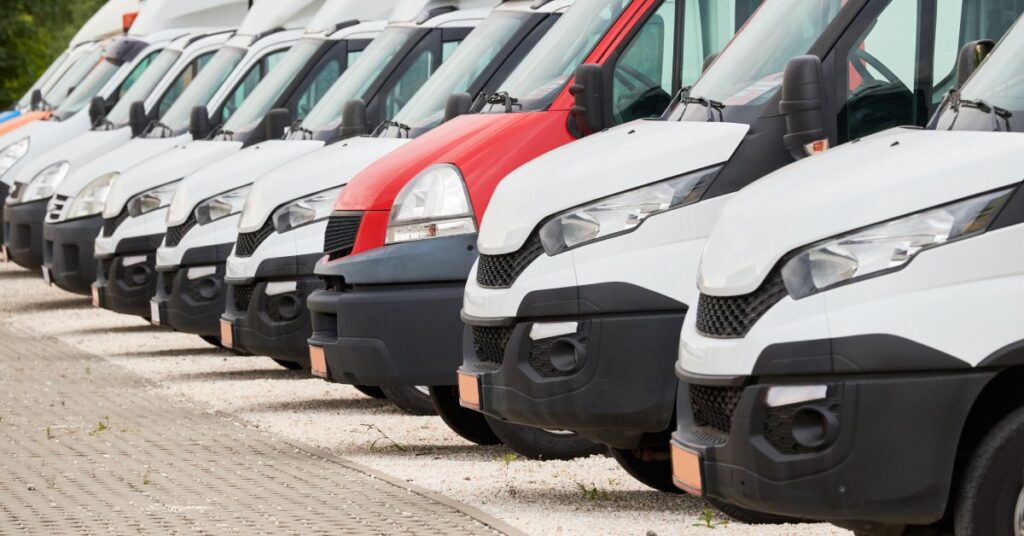 A row of parked commercial vans. They are all white, except for a single red van standing out among them on a paved lot.