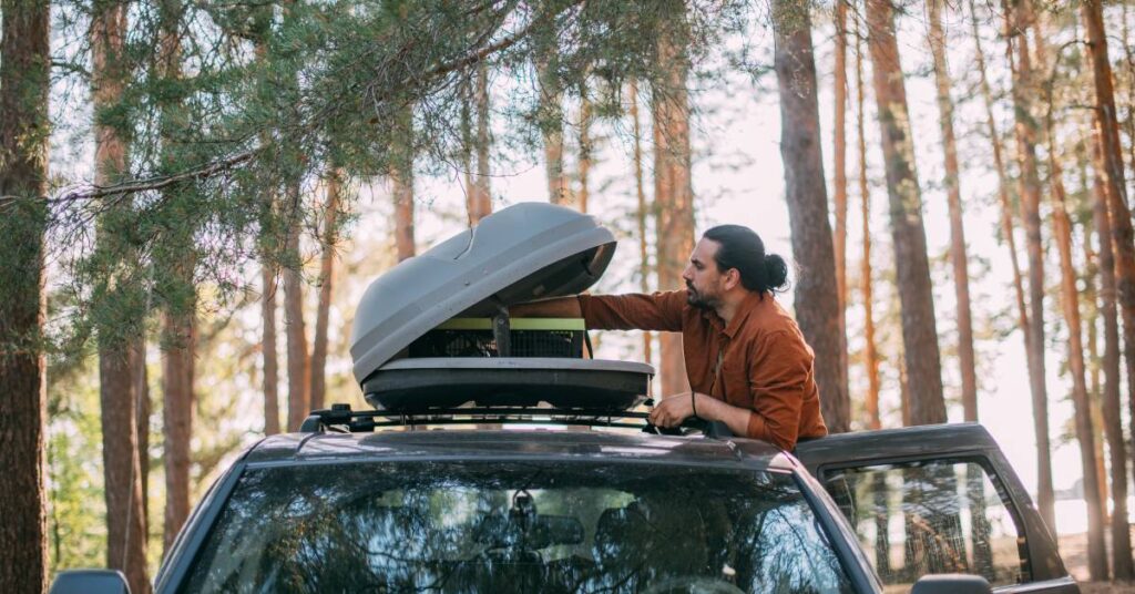 A young man opening the upper hard-shell trunk attached to his vehicle's roof rack in a pine forest on the lakeshore.