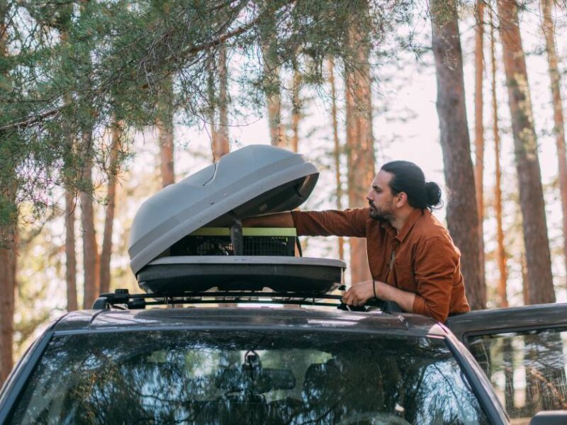 A young man opening the upper hard-shell trunk attached to his vehicle's roof rack in a pine forest on the lakeshore.