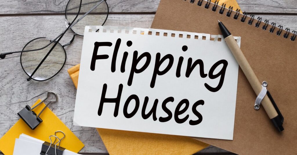 A cluttered wood desk featuring notebooks, a pair of glasses, post-it notes, and a torn page reading “Flipping Houses.”