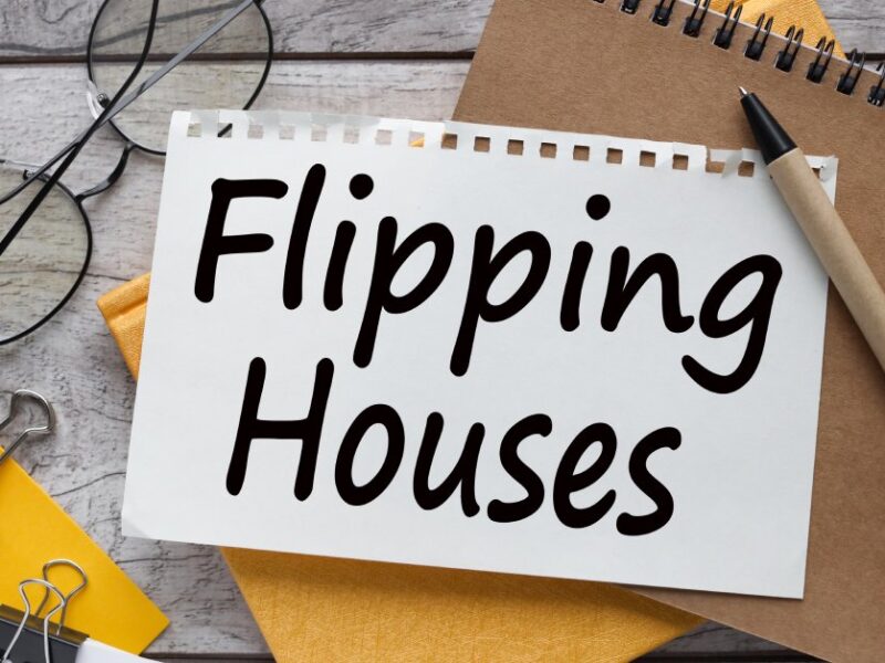 A cluttered wood desk featuring notebooks, a pair of glasses, post-it notes, and a torn page reading “Flipping Houses.”