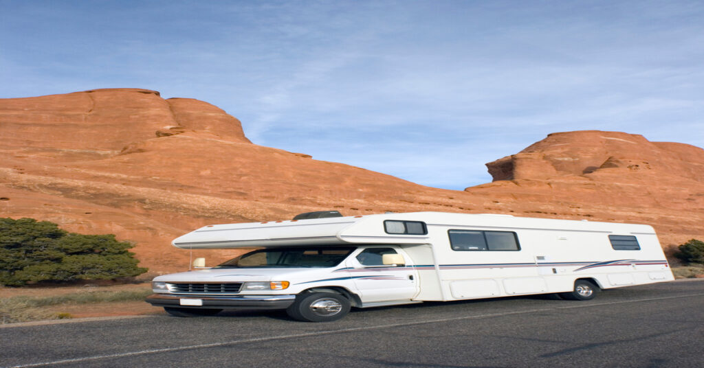 A white RV is traveling down the road. The road has large, orange rock formations next to it, and the sky is blue.