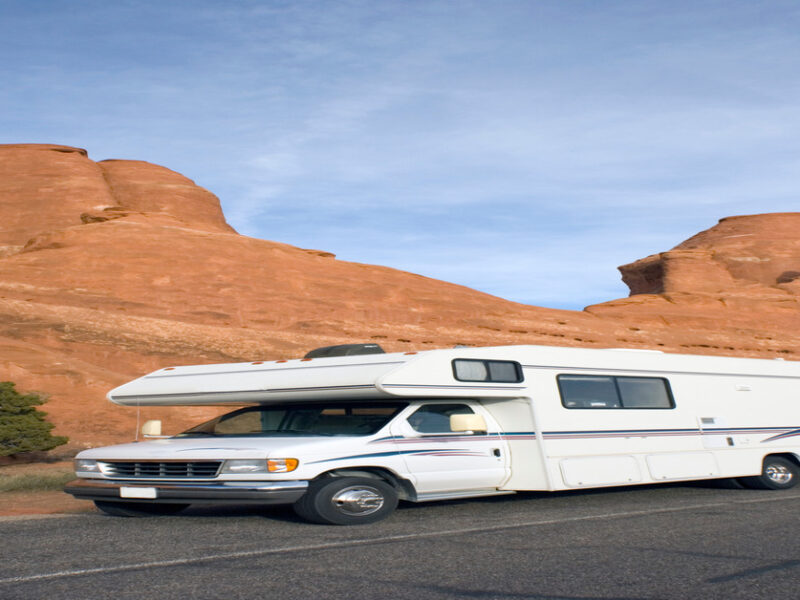 A white RV is traveling down the road. The road has large, orange rock formations next to it, and the sky is blue.