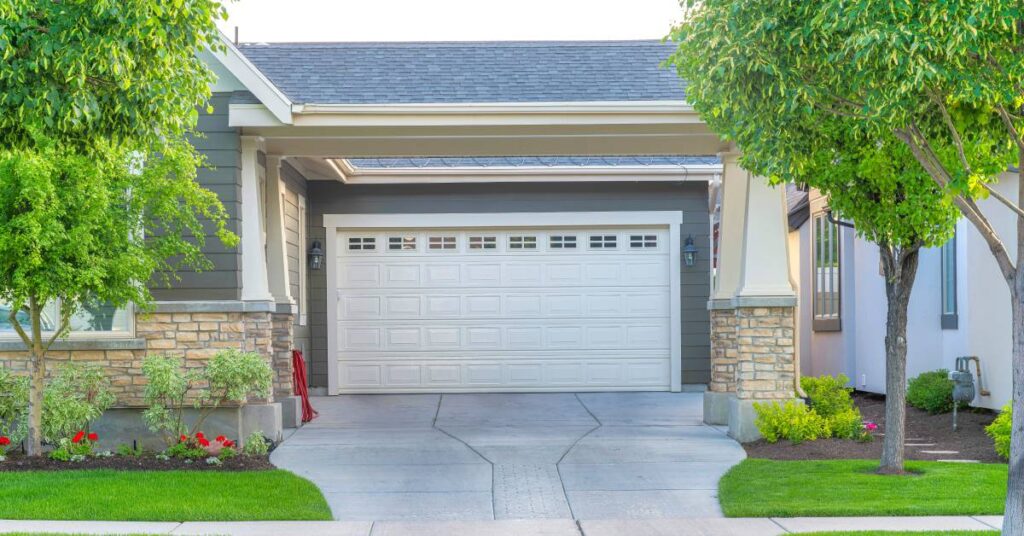 A concrete driveway that passes underneath a covered overhang that leads to the garage. There are some trees and plants nearby.