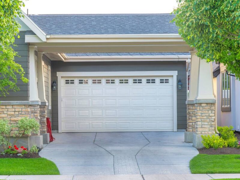 A concrete driveway that passes underneath a covered overhang that leads to the garage. There are some trees and plants nearby.